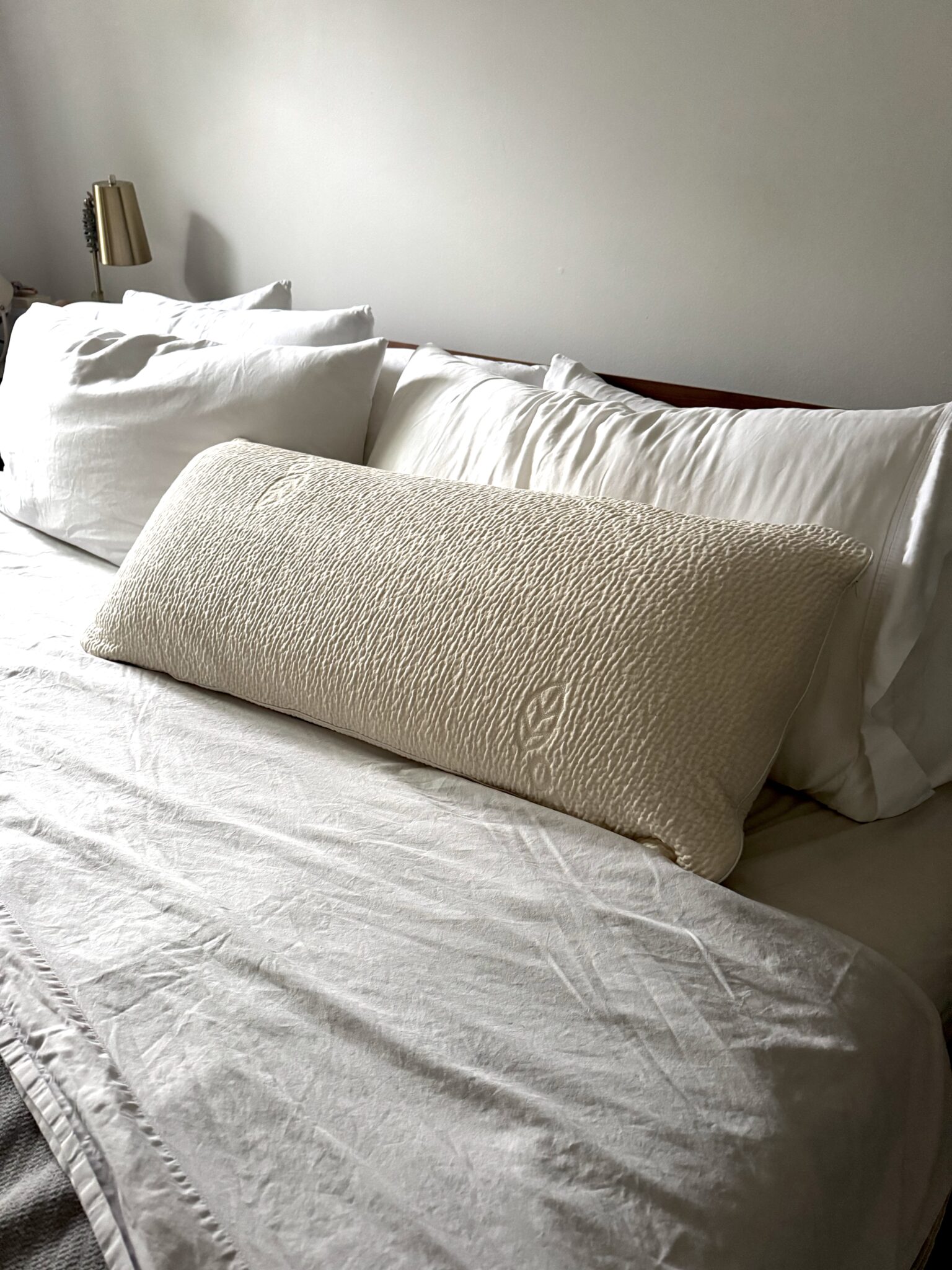 A neatly made bed with white sheets and pillows, featuring a large textured beige body pillow in front of two standard pillows.
