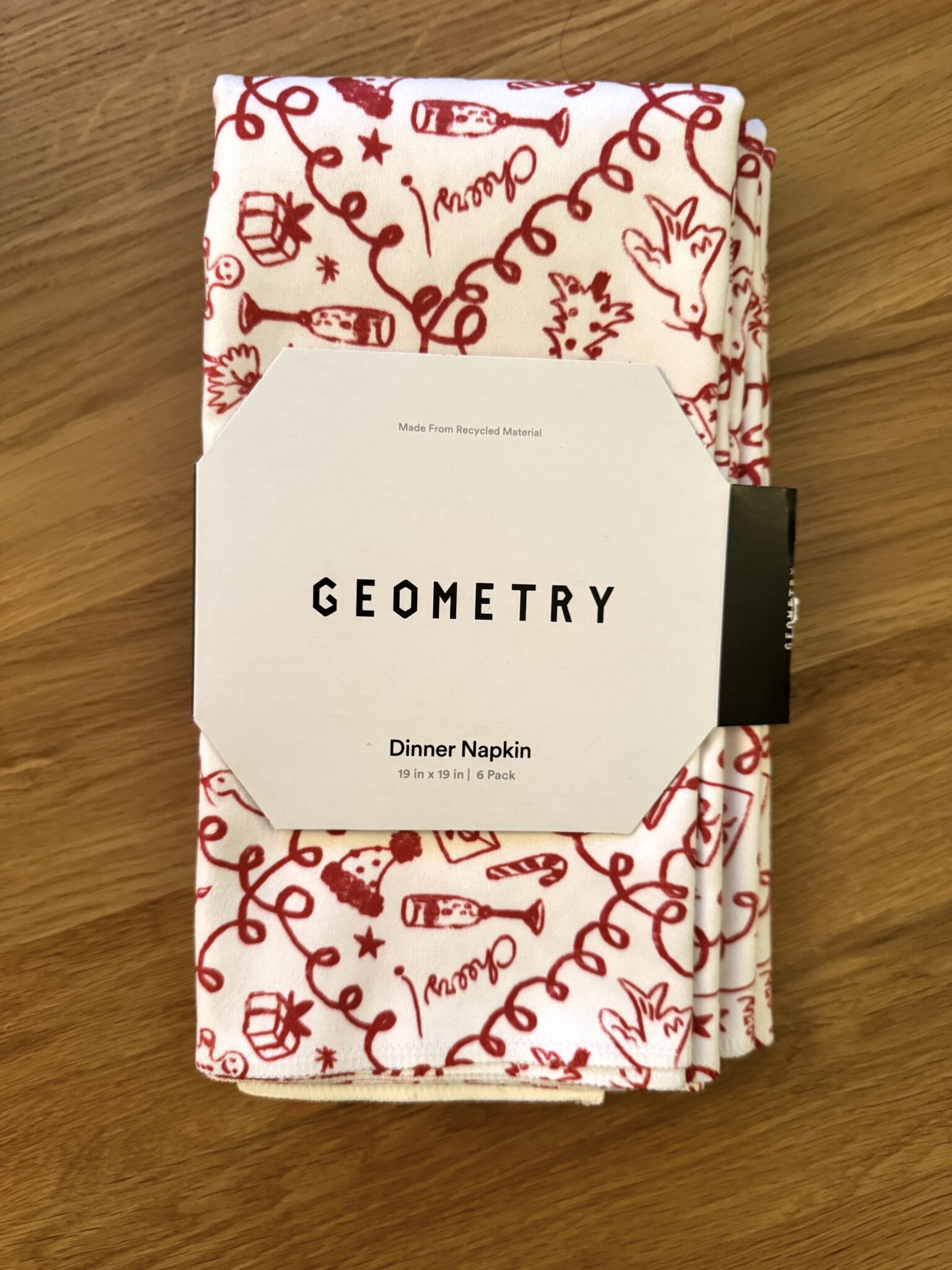 A folded white dinner napkin with red illustrations, labeled "GEOMETRY Dinner Napkin" in a 6-pack, sits on a wooden surface.