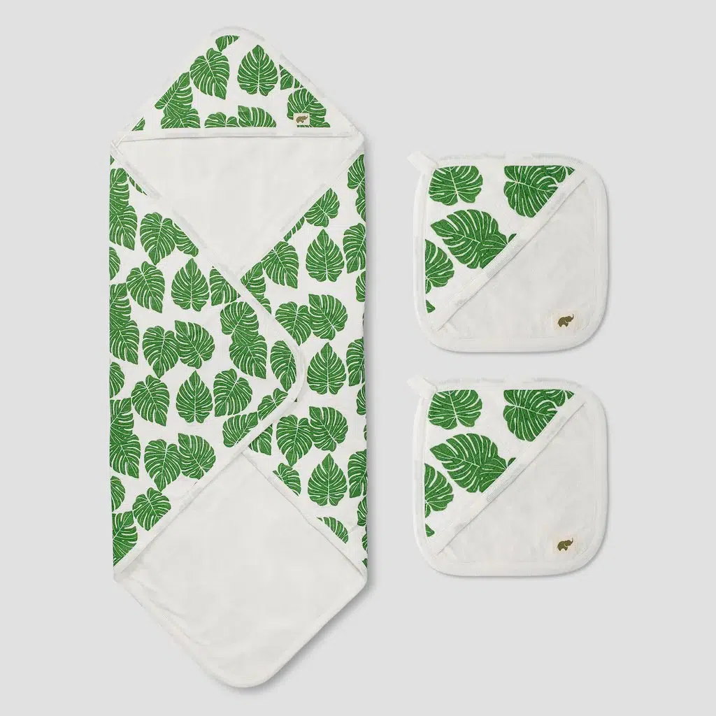 A white hooded baby towel and two matching washcloths with green monstera leaf prints are displayed on a light background.