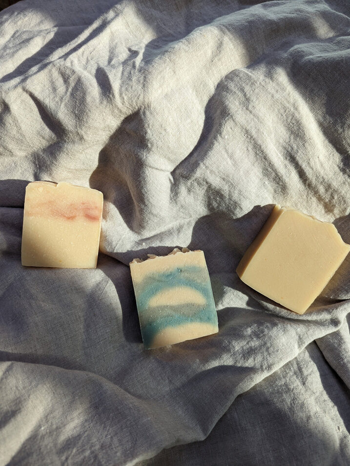 Three rectangular bars of handmade soap, one with a pink swirl, one with blue and white swirls, and one plain, arranged on a wrinkled light-colored fabric.