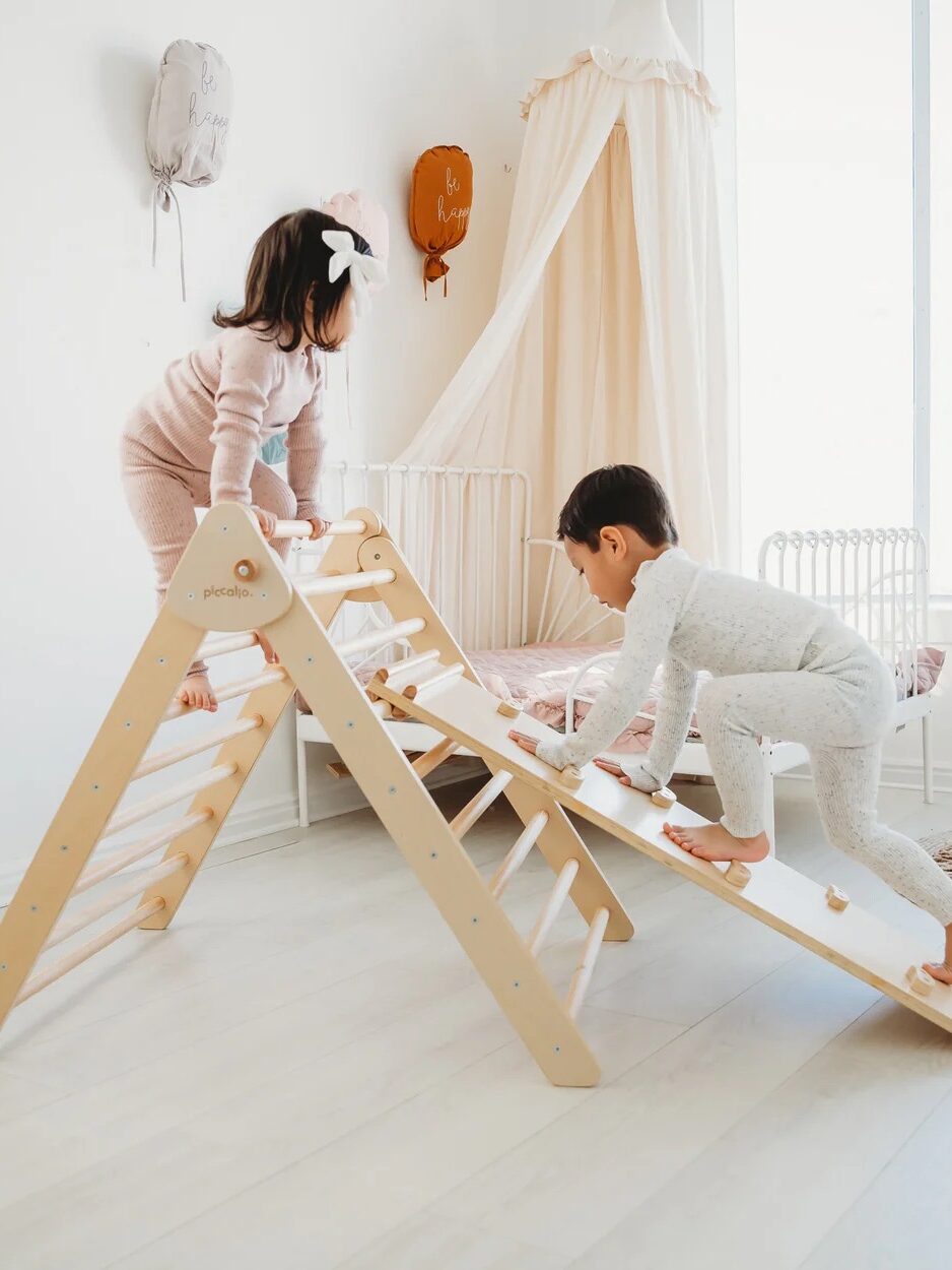 Two children in pajamas climb and play on a wooden indoor climbing frame and ramp in a bright, minimalistic bedroom.