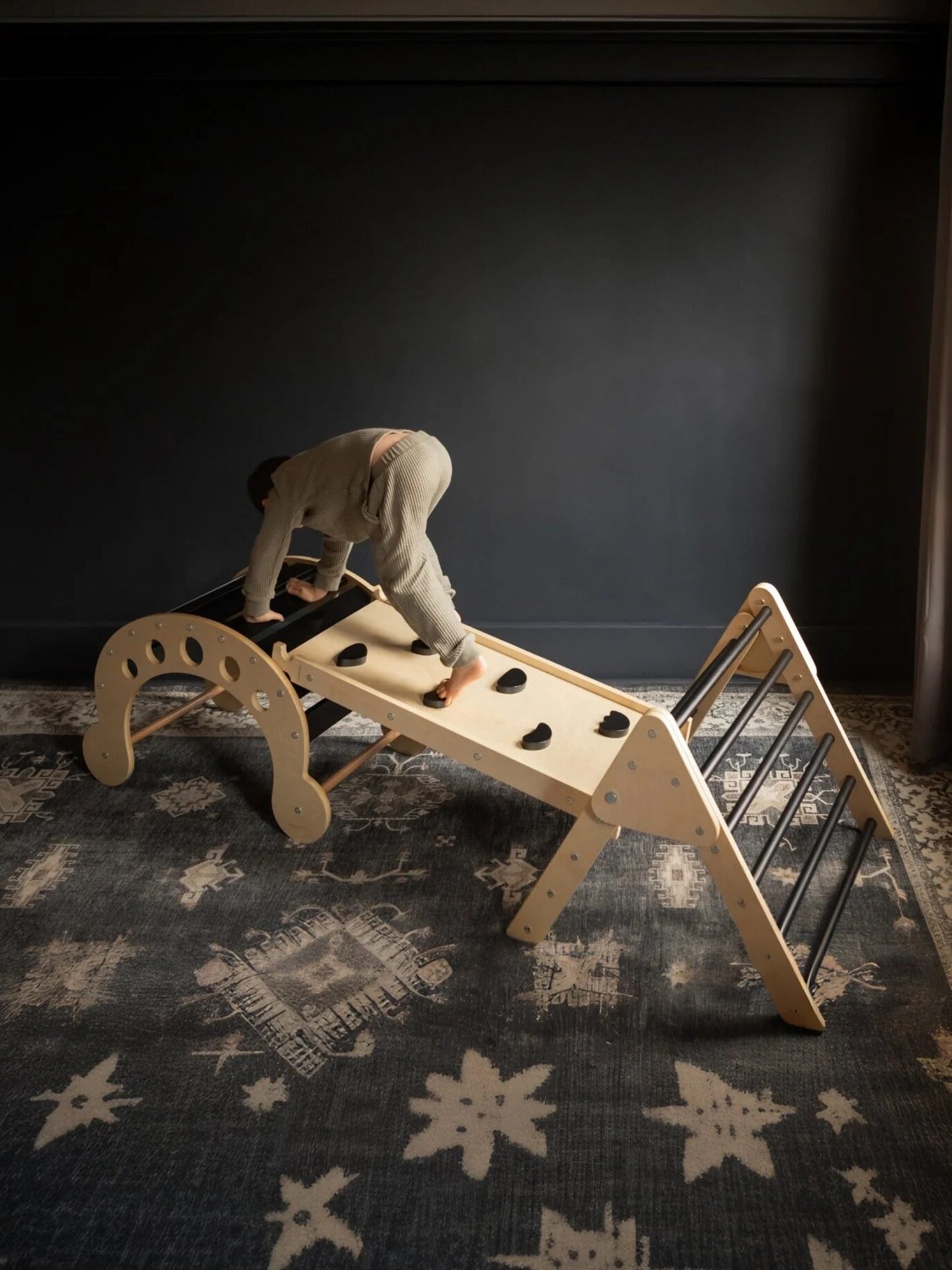 Child climbing on a wooden indoor play structure with ladder, bridge, and climbing wall features, set on a patterned rug against a dark wall.