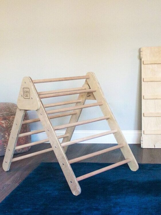A wooden climbing triangle and two flat panels are placed on a blue rug in a room with light walls and two patterned poufs in the corner.