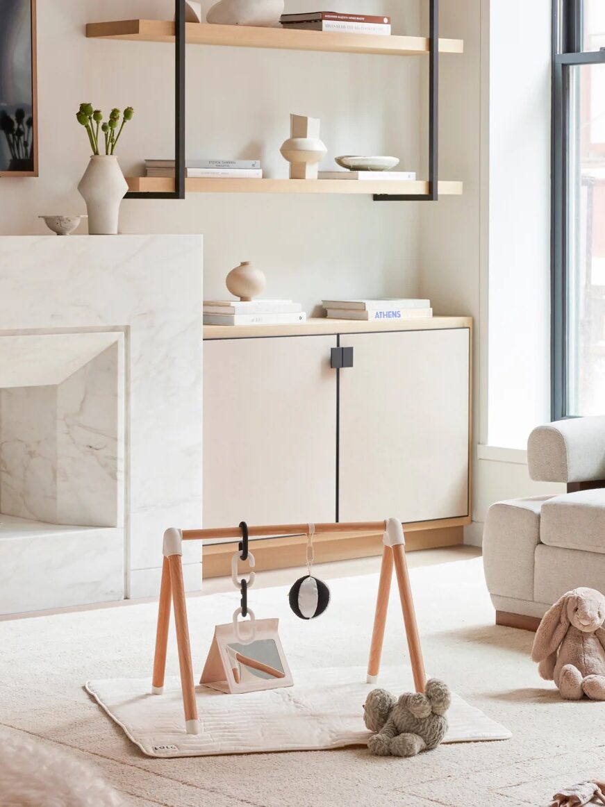 A modern living room with a baby play gym on a white mat, a plush toy, shelves with decor, and a cabinet against a light-colored wall.