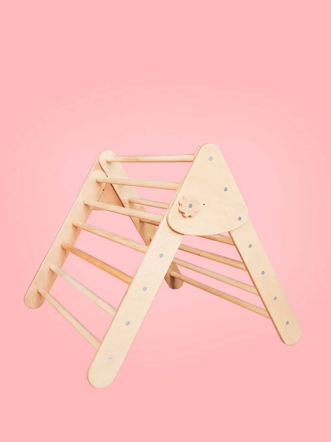 A wooden climbing triangle for children is placed against a solid pink background.