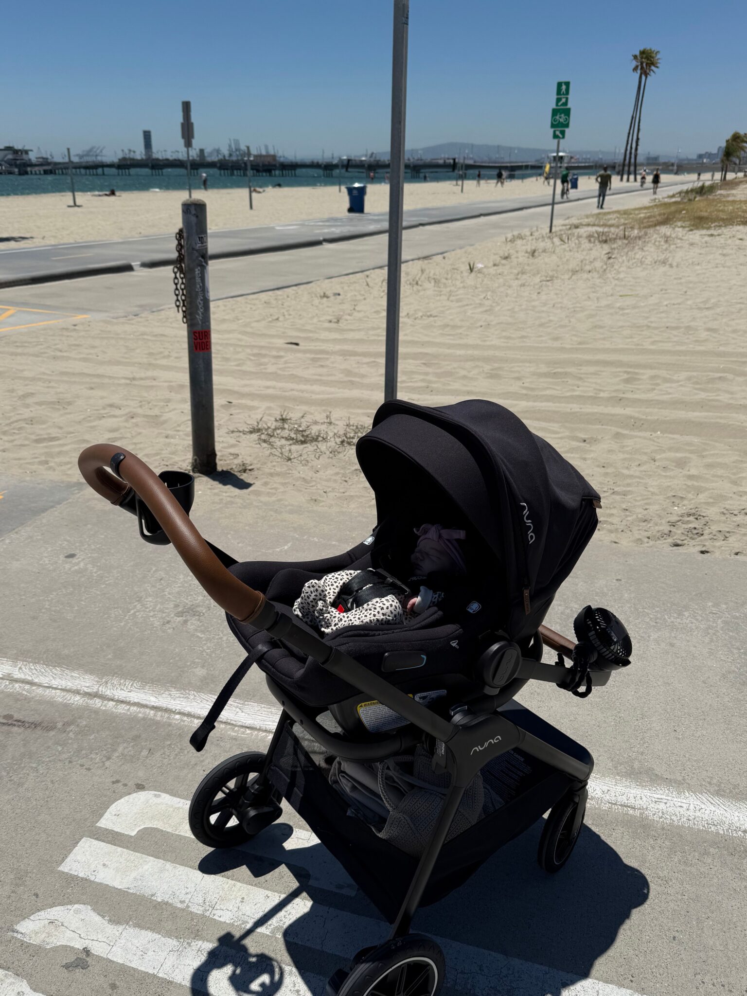 A black baby stroller is parked on a paved path by a sandy beach under a clear blue sky.