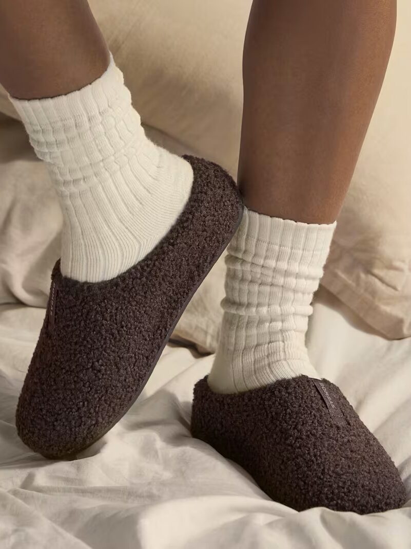 A person wearing white socks and dark brown slippers stands on a bed with cream-colored bedding.