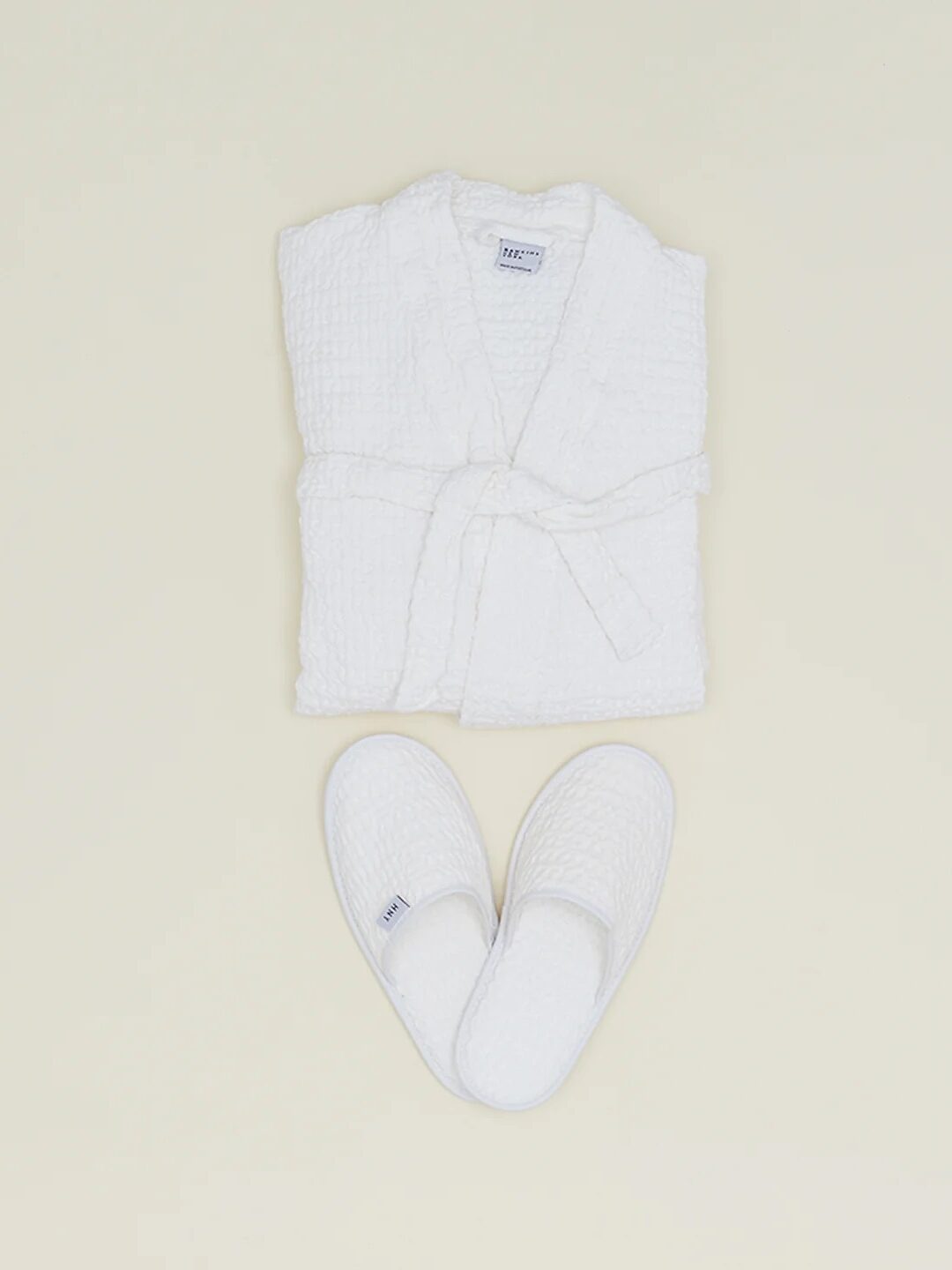 A folded white bathrobe with a belt rests neatly above a pair of cozy slippers on a plain light background.