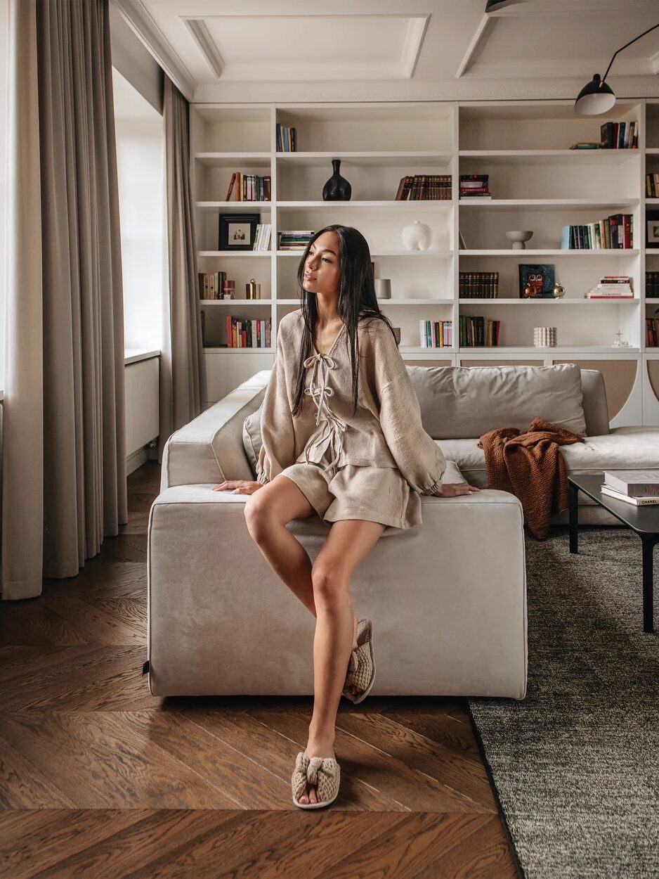 Woman in beige loungewear and slippers sits on a sofa in a modern living room with bookshelves, a coffee table, and large windows.