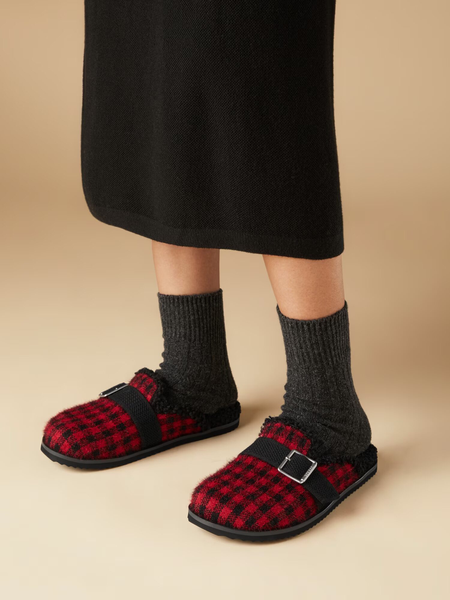 Person wearing dark socks and a black skirt paired with red-and-black checkered slippers featuring a buckle strap, standing on a neutral beige background. These cozy slippers add a stylish touch to the casual look.