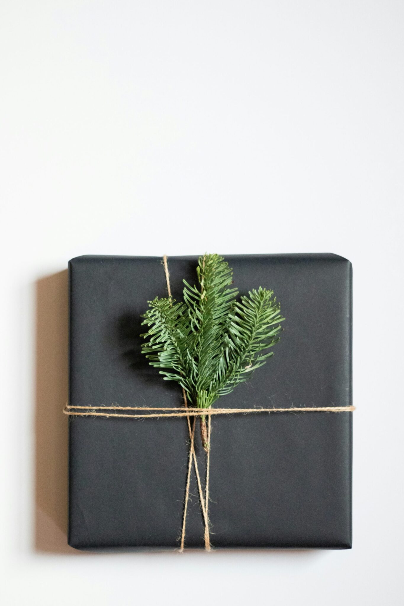 A square gift wrapped in black paper is tied with twine and decorated with a small sprig of evergreen on a plain white background.