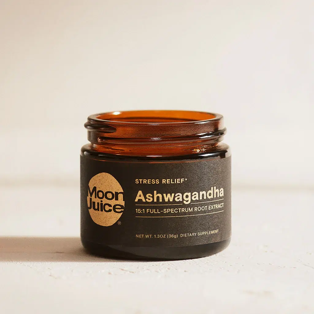 A brown glass jar labeled "Moon Juice Ashwagandha 15:1 Full-Spectrum Root Extract" sits on a light surface. The label notes it is a dietary supplement for stress relief.