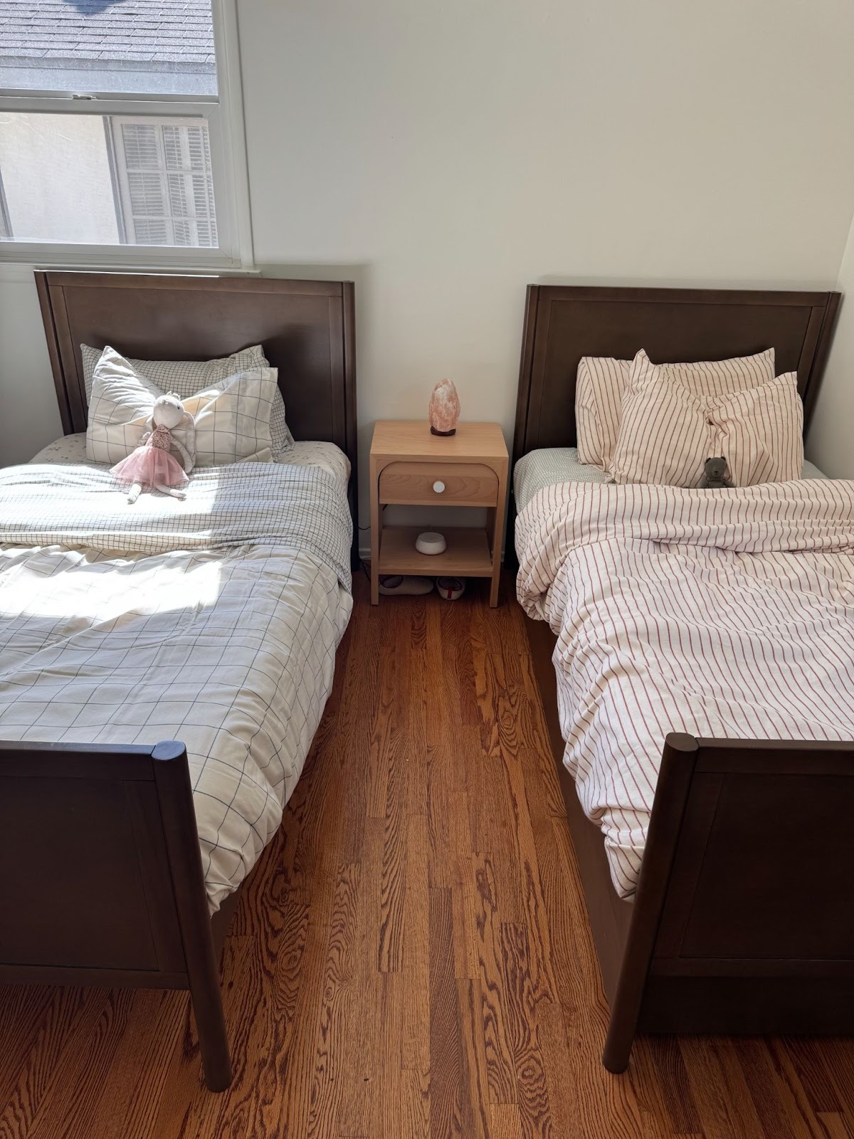 Two twin beds with wooden frames are placed side by side, separated by a small nightstand, in a sunlit bedroom with hardwood floors.